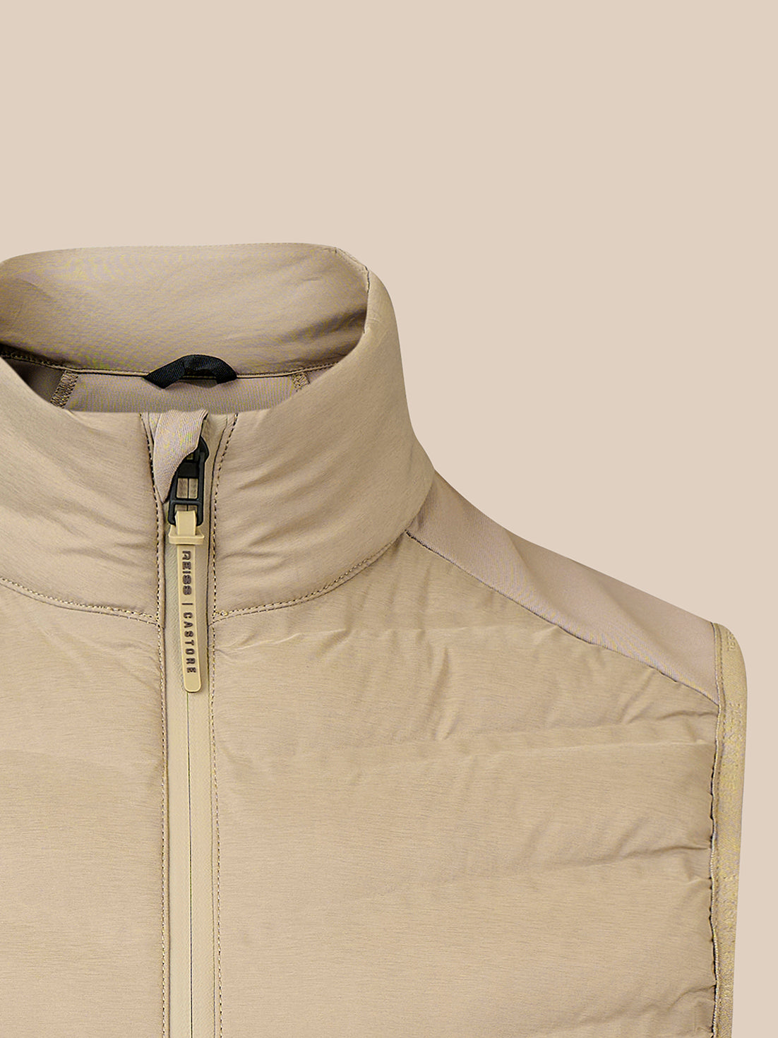 Close-up of a beige puffer vest with a high stand-up collar and a gold-toned zipper pull, shown against a solid beige background.