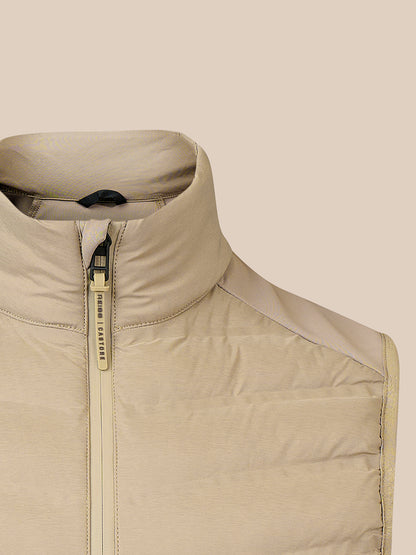Close-up of a beige puffer vest with a high stand-up collar and a gold-toned zipper pull, shown against a solid beige background.