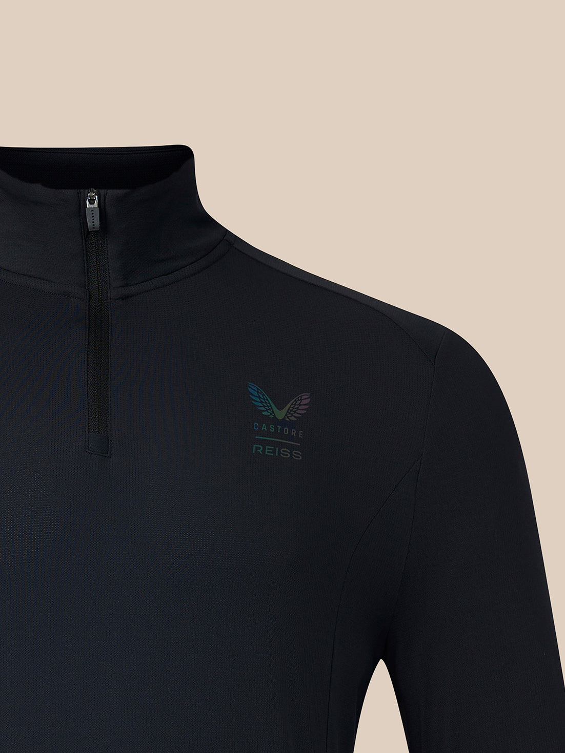 Close-up of a black long-sleeve quarter-zip athletic top with a small multicolor winged logo and the text 'CASTORE REISS' on the left chest, shown against a solid beige background.