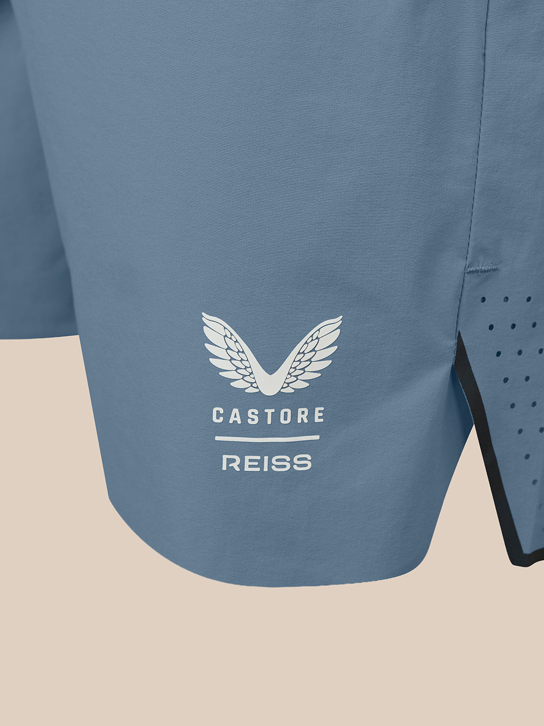 Close-up of a blue athletic shorts leg featuring a white Castore Reiss wing logo on the front, with a side vent showing black lining and perforated detailing, set against a solid beige background.
