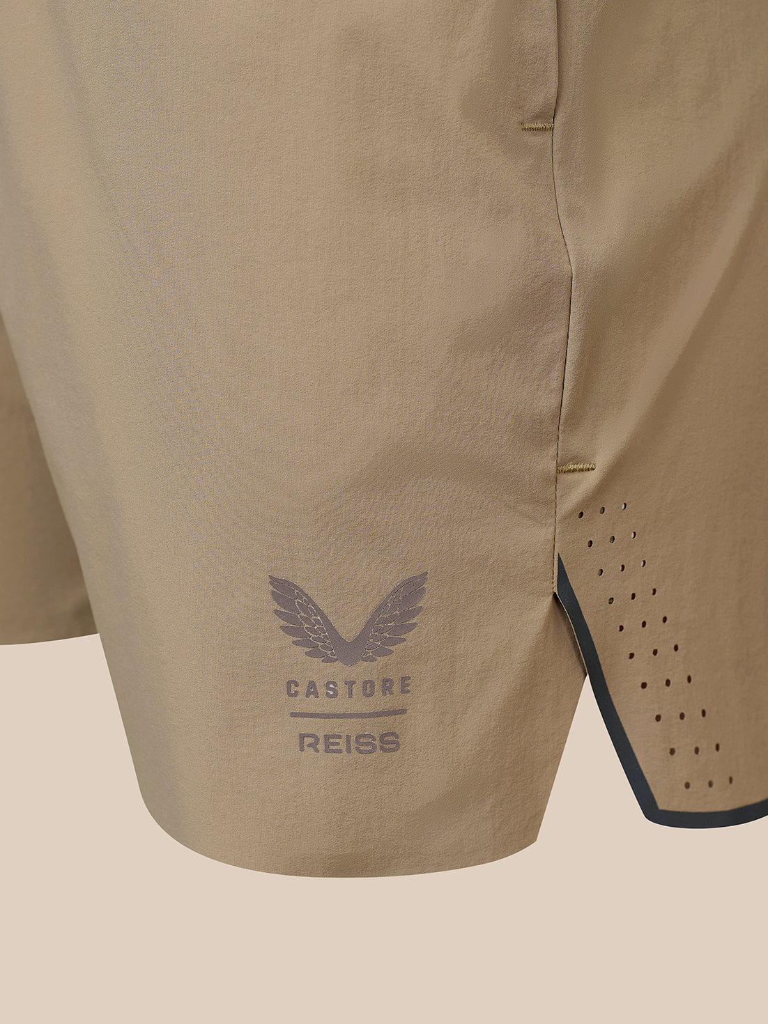 Close-up of beige Castore Reiss athletic shorts showing the side seam, perforated side panel, and a dark gray wing logo with brand text.