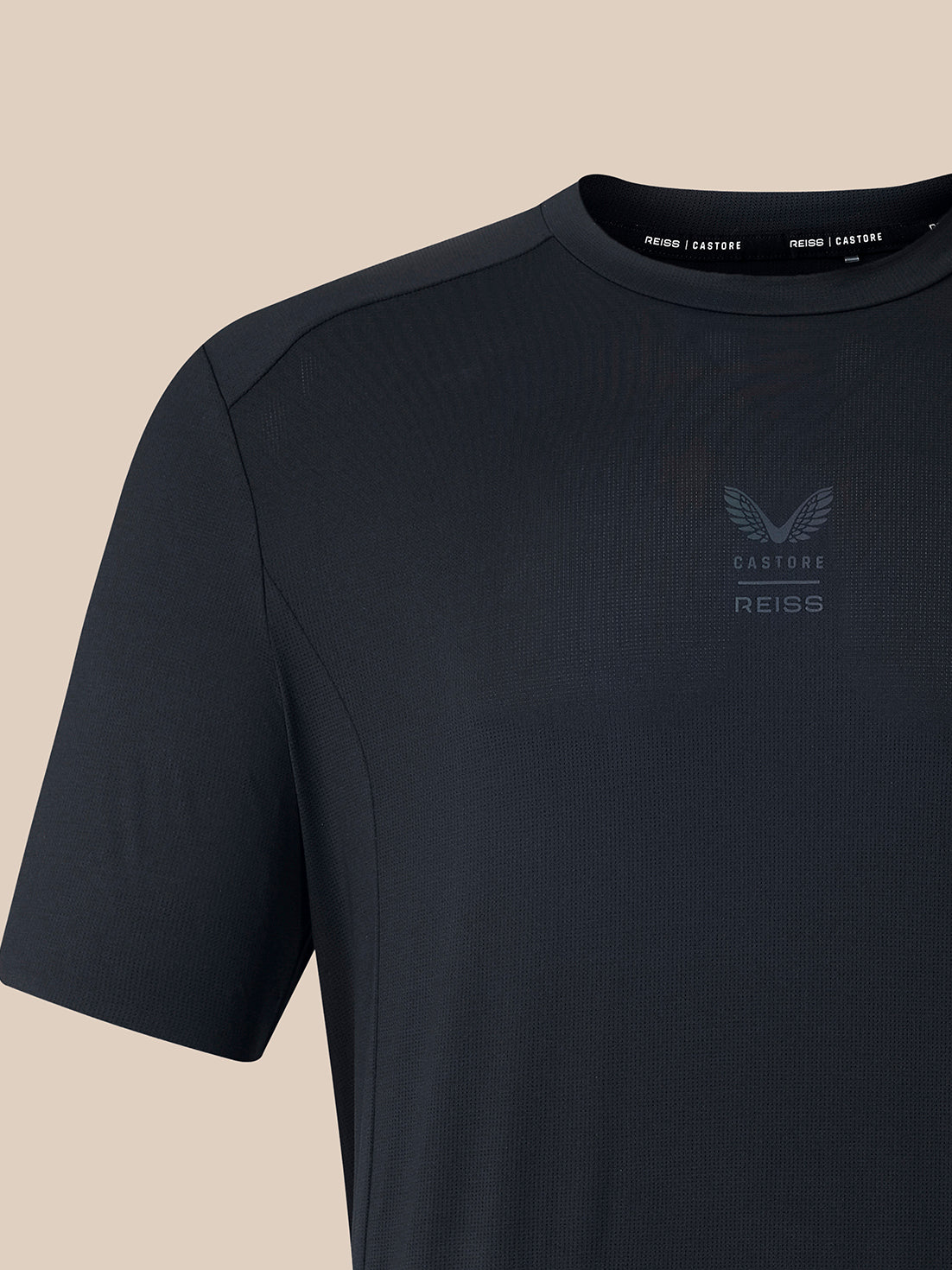 A close-up of a dark charcoal gray Castore Reiss athletic t-shirt on a solid beige background.