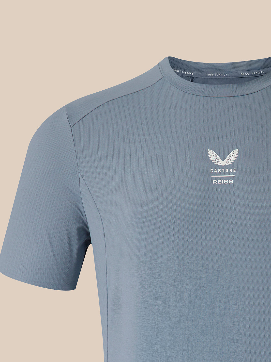 A close-up of a light blue athletic t-shirt with a white winged logo and the text 'CASTORE REISS' on the left chest, set against a solid beige background.