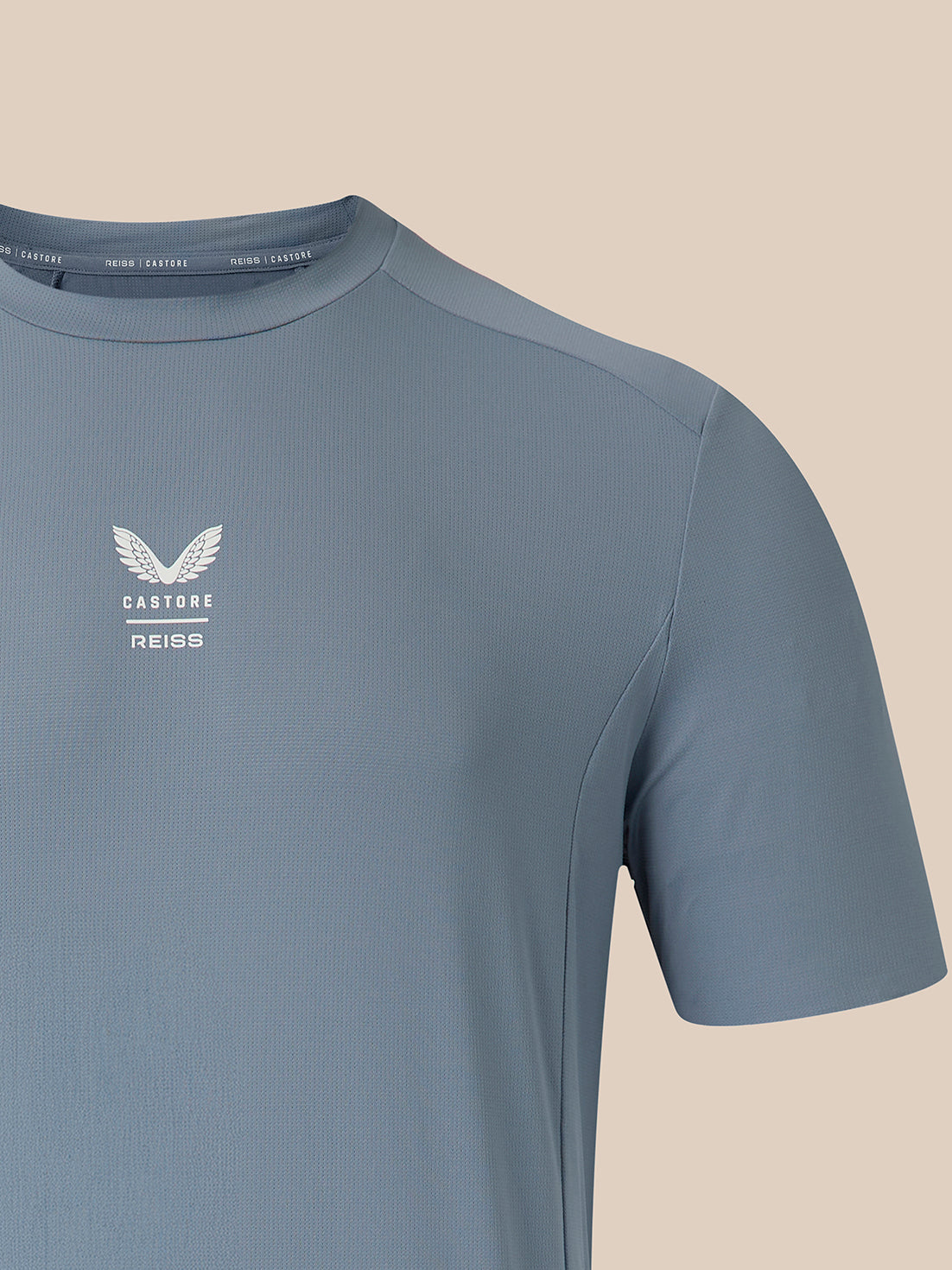 A close-up of a slate blue athletic t-shirt featuring a white Castore Reiss wing logo on the left chest, set against a solid beige background.