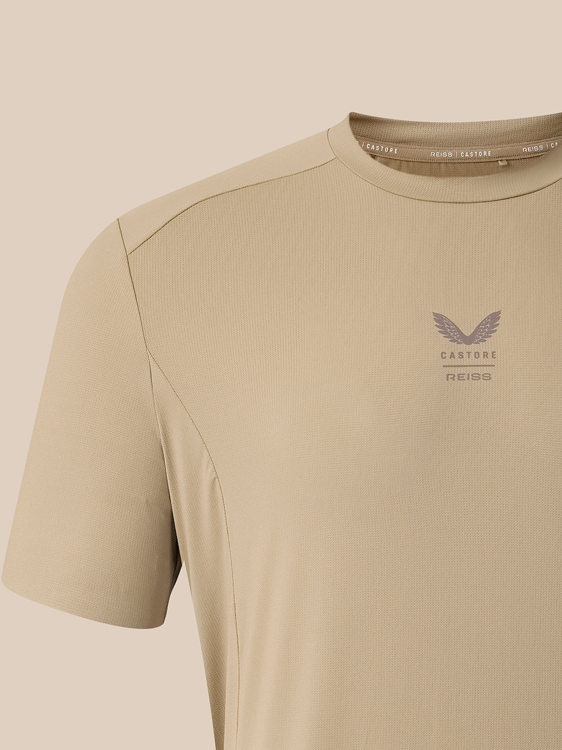 Close-up of a beige short-sleeve athletic t-shirt with a subtle winged logo and the text 'CASTORE REISS' printed on the left chest, set against a matching beige background.