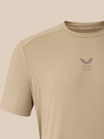 Close-up of a beige short-sleeve athletic t-shirt with a subtle winged logo and the text 'CASTORE REISS' printed on the left chest, set against a matching beige background.