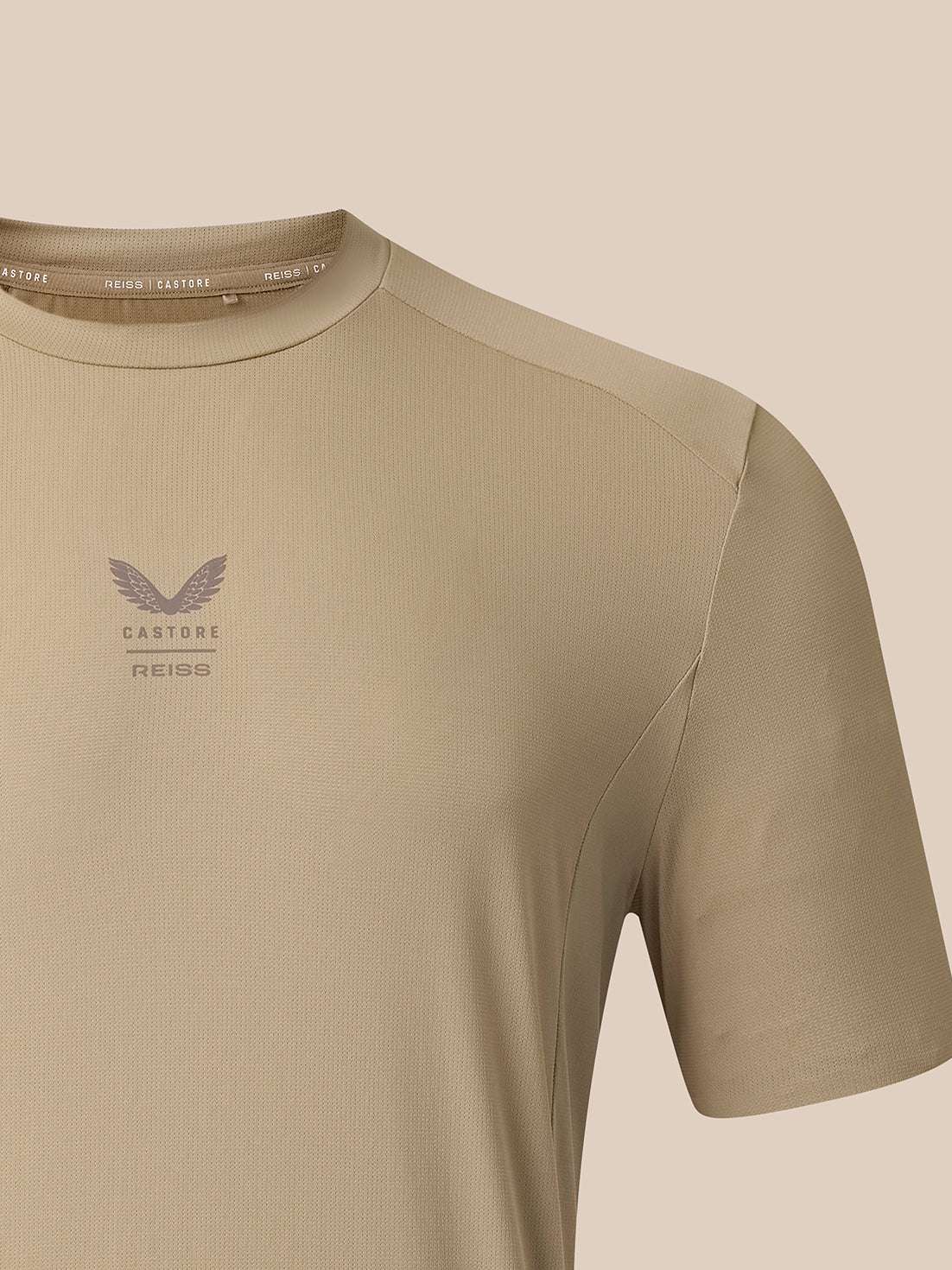 Close-up of a short-sleeve athletic t-shirt in a warm beige/khaki color, featuring a subtle winged logo and the text 'CASTORE REISS' on the left chest, set against a solid beige background.