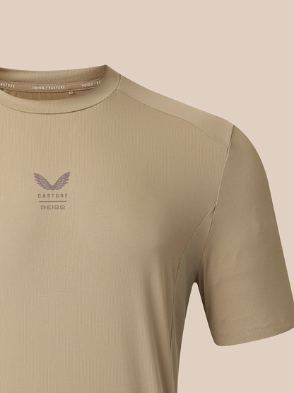 Close-up of a short-sleeve athletic t-shirt in a warm beige/khaki color, featuring a subtle winged logo and the text 'CASTORE REISS' on the left chest, set against a solid beige background.
