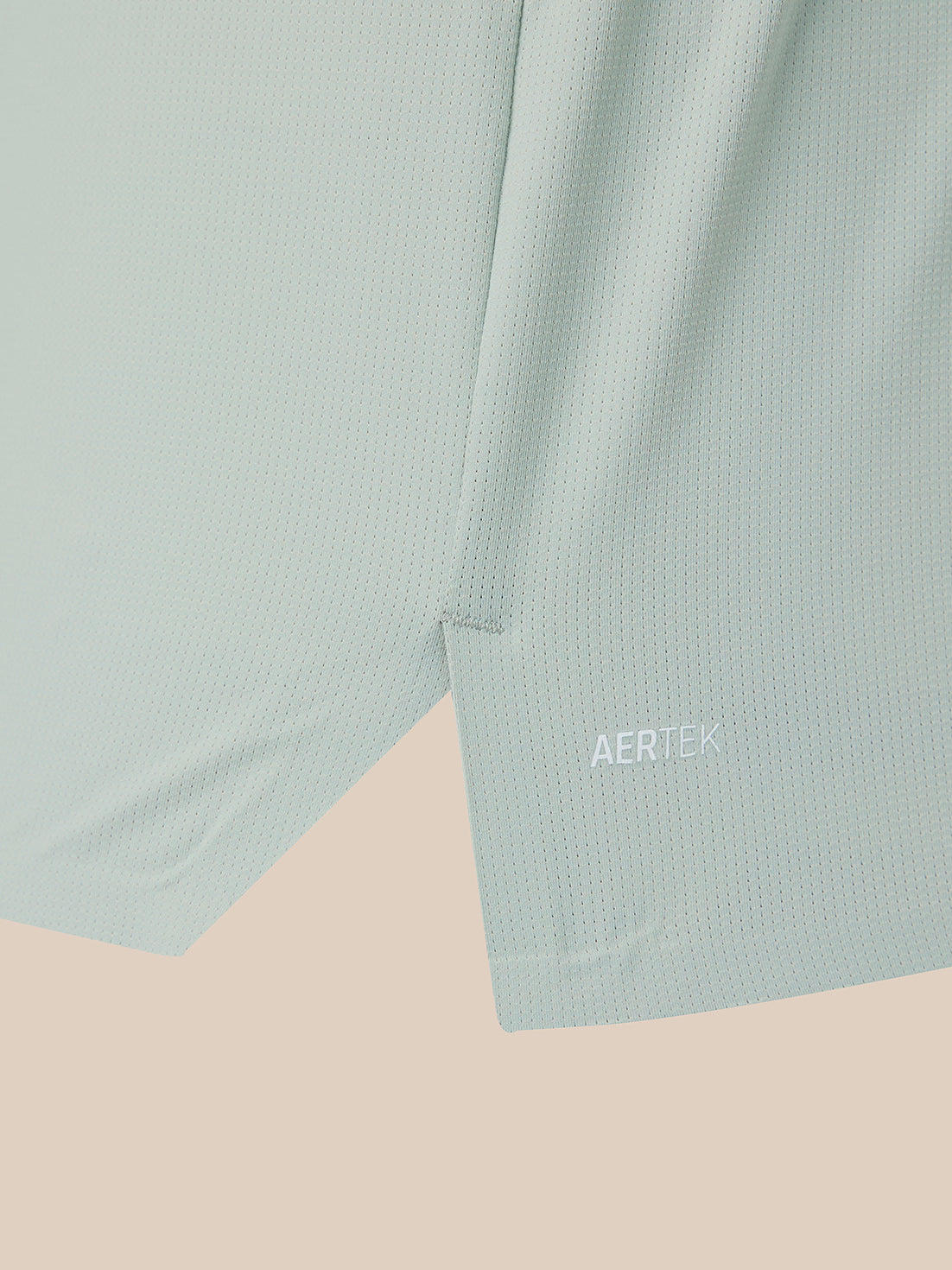 Close-up of a light mint green athletic fabric with a subtle textured mesh pattern, featuring a small white AERTEK logo near the lower hem, set against a solid pale peach background.