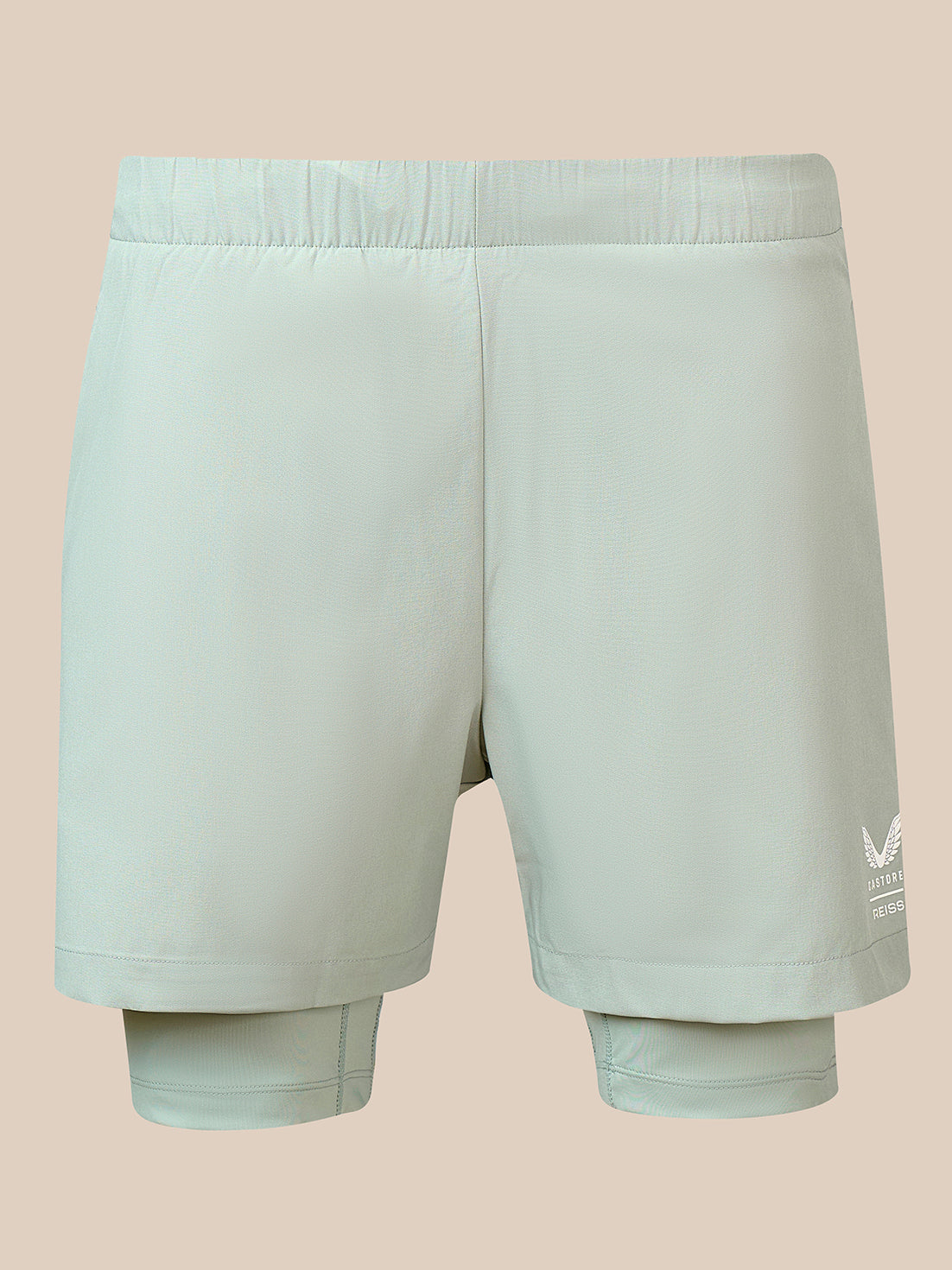 A pair of light sage green athletic shorts shown from the back, featuring a built-in compression liner at the hem with a white 'STOP' logo on the left leg, set against a solid beige background.