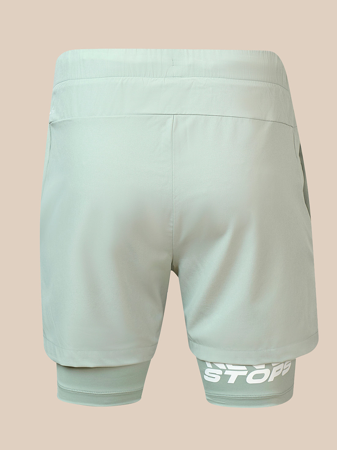 A pair of light sage green athletic shorts shown from the back, featuring a built-in compression liner at the hem with a white 'STOP' logo on the left leg, set against a solid beige background.
