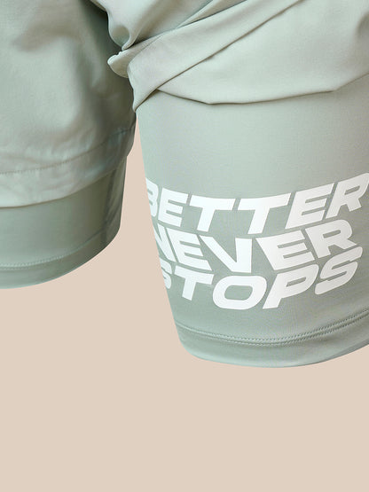 Close-up of sage green athletic shorts with a large white graphic text reading 'BETTER NEVER STOPS' on the thigh, set against a solid pale beige background.