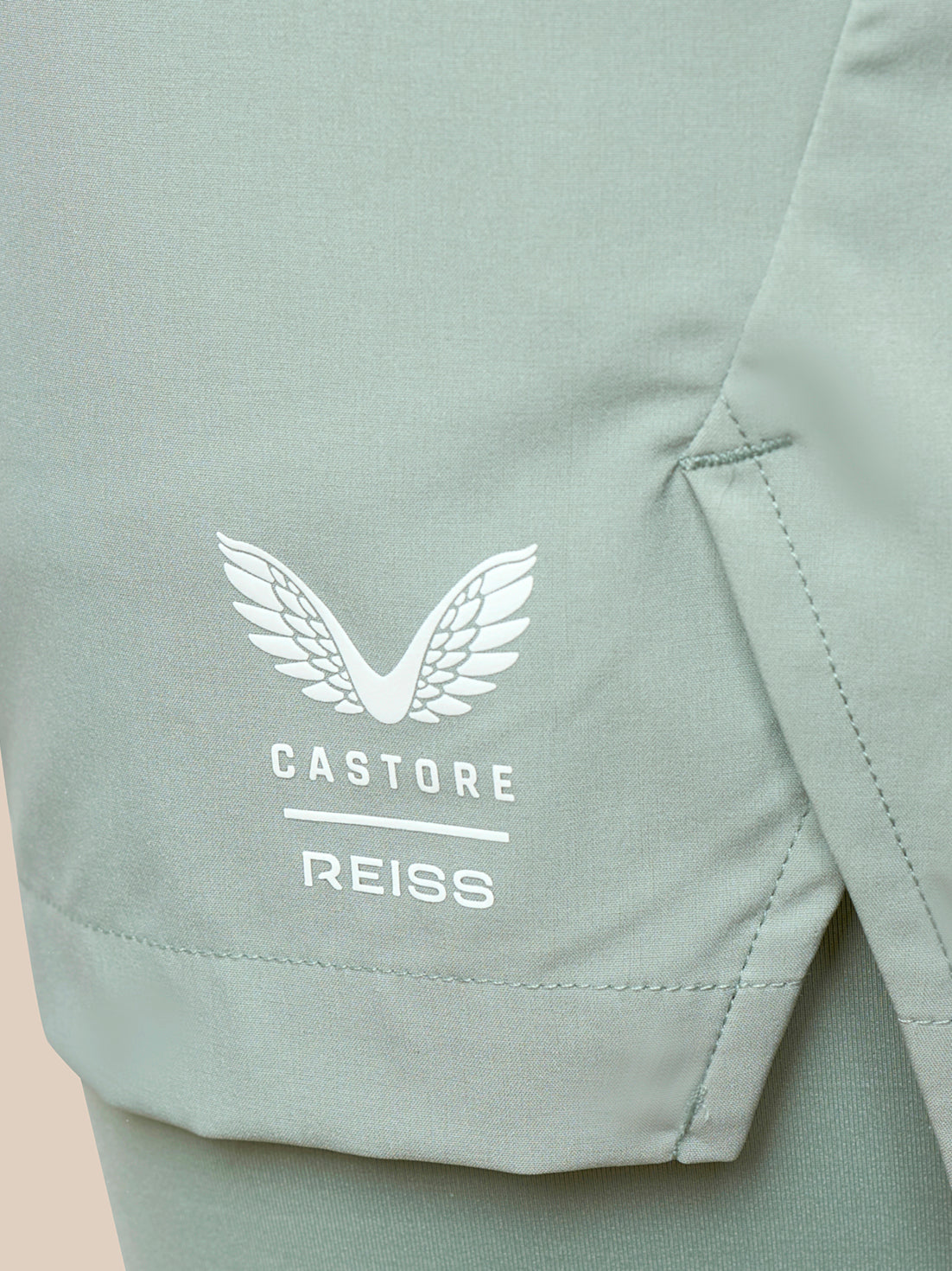 Close-up of a sage green athletic garment showing a white embroidered wing logo and the text 'CASTORE REISS' on the side.