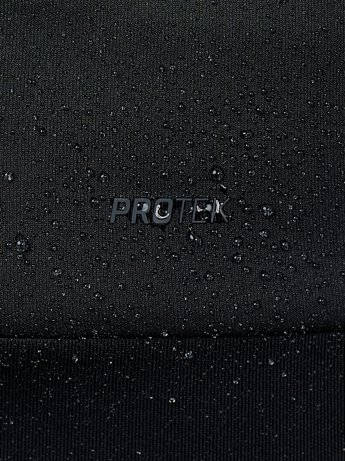 Close-up of a black textured fabric with water droplets beaded on the surface and a small metallic 'PROTEK' logo in the center.