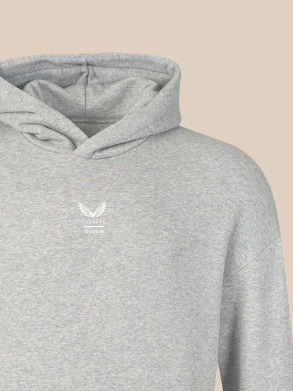 A close-up of a light heather gray hoodie with a small white embroidered winged logo and the word 'CASTORE' on the left chest, shown against a solid beige background.