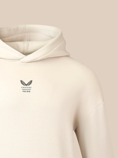 Close-up of a cream-colored hooded sweatshirt with a small black winged logo and the brand name 'CASTORE' printed on the left chest, set against a solid beige background.
