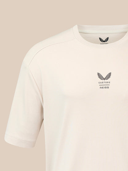 A close-up of a light cream-colored Castore Reiss short-sleeve t-shirt, showing the inner neck label and the chest logo on a solid beige background.