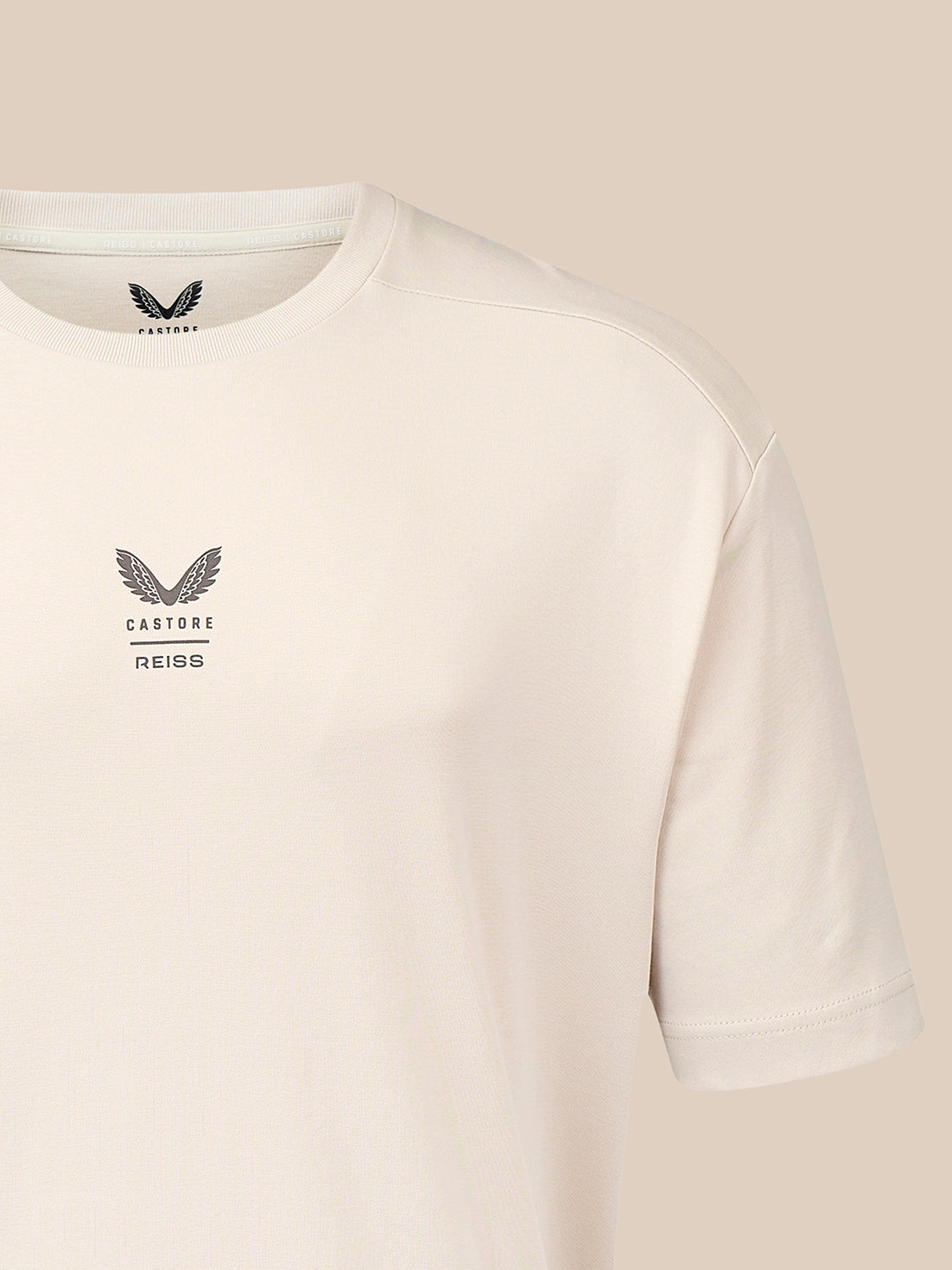 A close-up of a light cream-colored Castore Reiss t-shirt, showing the left chest logo and the inner neck label, set against a solid beige background.