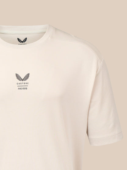 A close-up of a light cream-colored Castore Reiss t-shirt, showing the left chest logo and the inner neck label, set against a solid beige background.