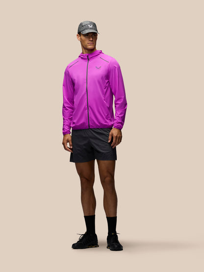 A male model wearing a bright purple athletic zip-up jacket, dark gray running shorts, a gray cap, black socks, and black running shoes, standing against a solid beige background.