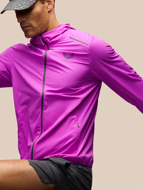 A man wearing a bright magenta-purple athletic zip-up jacket with a small winged logo on the left chest that reads 'CASTORE'. The jacket has black zipper details and is paired with black running shorts against a solid beige background.