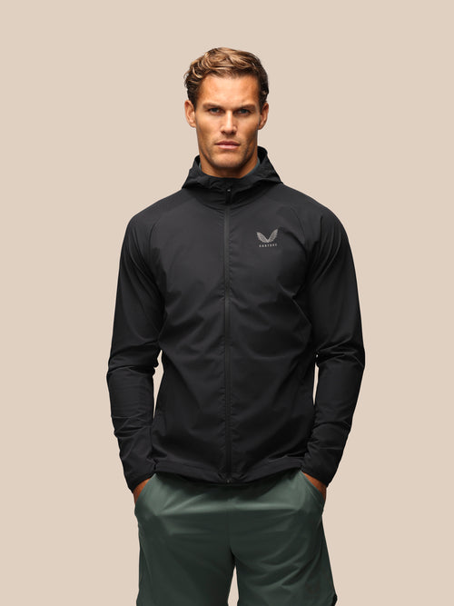 A man wearing a black hooded zip-up jacket with a small gray wing logo on the left chest, paired with dark green pants, standing against a solid beige background.