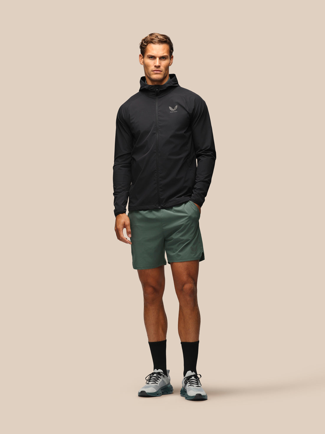 A male model stands against a solid beige studio background wearing a black lightweight zip-up hooded jacket, olive green athletic shorts, black socks, and gray running shoes.
