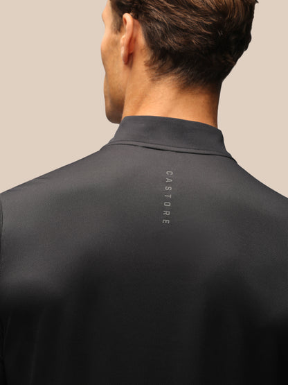 Close-up back view of a man wearing a dark charcoal gray mock neck shirt with the brand name 'CASTORE' printed vertically on the upper back, against a solid beige background.