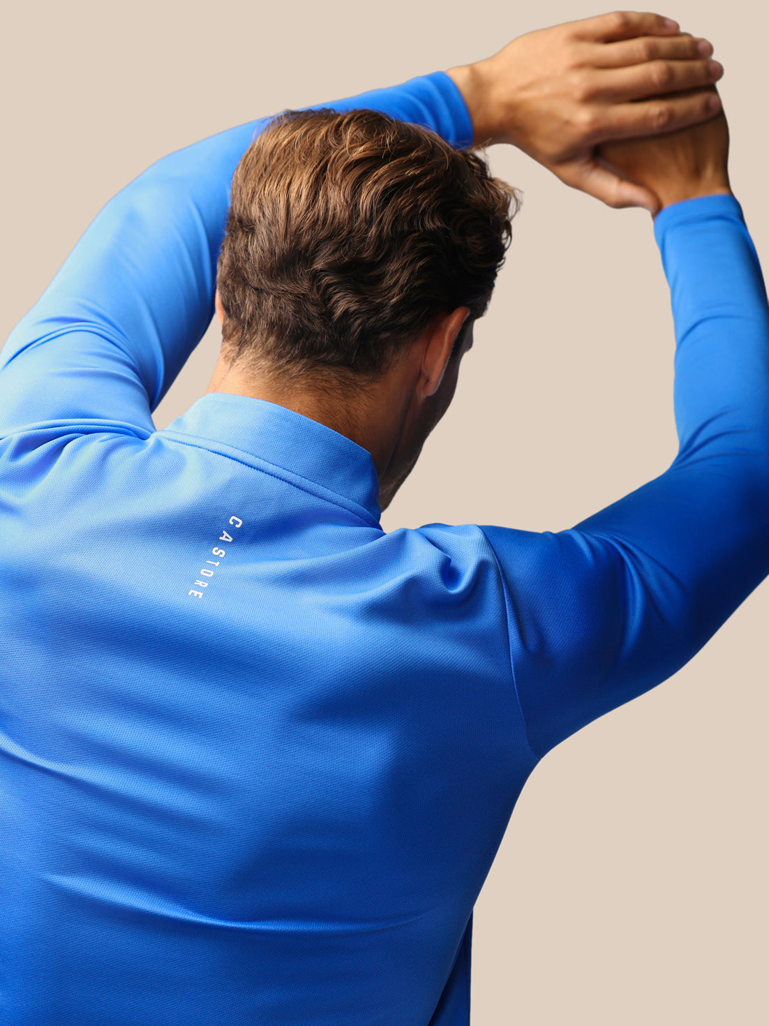 A man wearing a bright blue long-sleeve athletic shirt, shown from behind with his arms raised, on a plain beige background. The brand name 'CASTORE' is printed vertically on the back of the shirt.