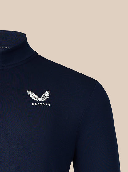 Close-up of a navy blue mock neck sweater with a white embroidered winged logo and the brand name 'CASTORE' on the left chest, set against a solid beige background.