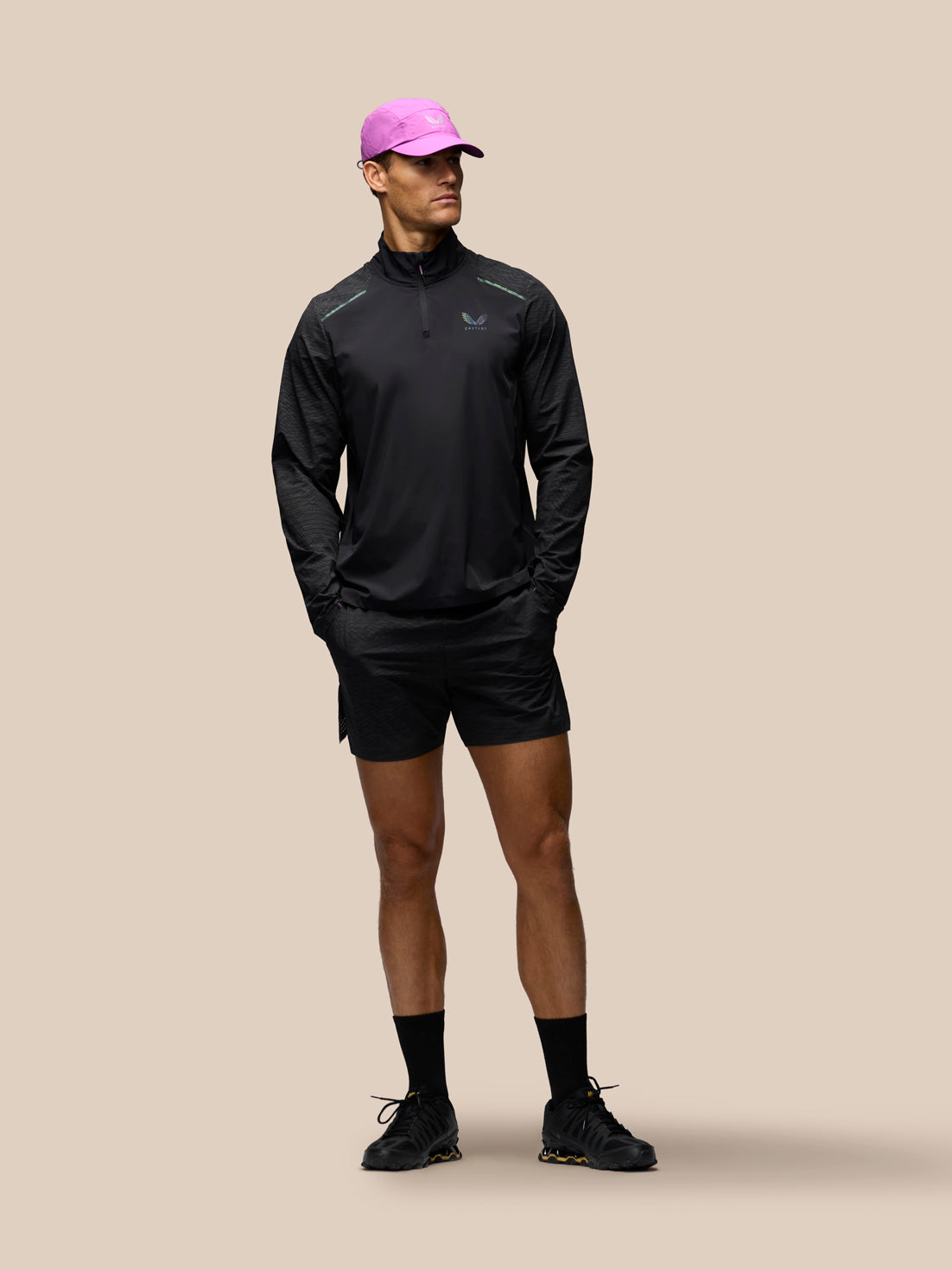 A male model wearing a black long-sleeve athletic quarter-zip pullover, black running shorts, black socks, and black running shoes, paired with a bright pink cap, standing against a solid beige background.