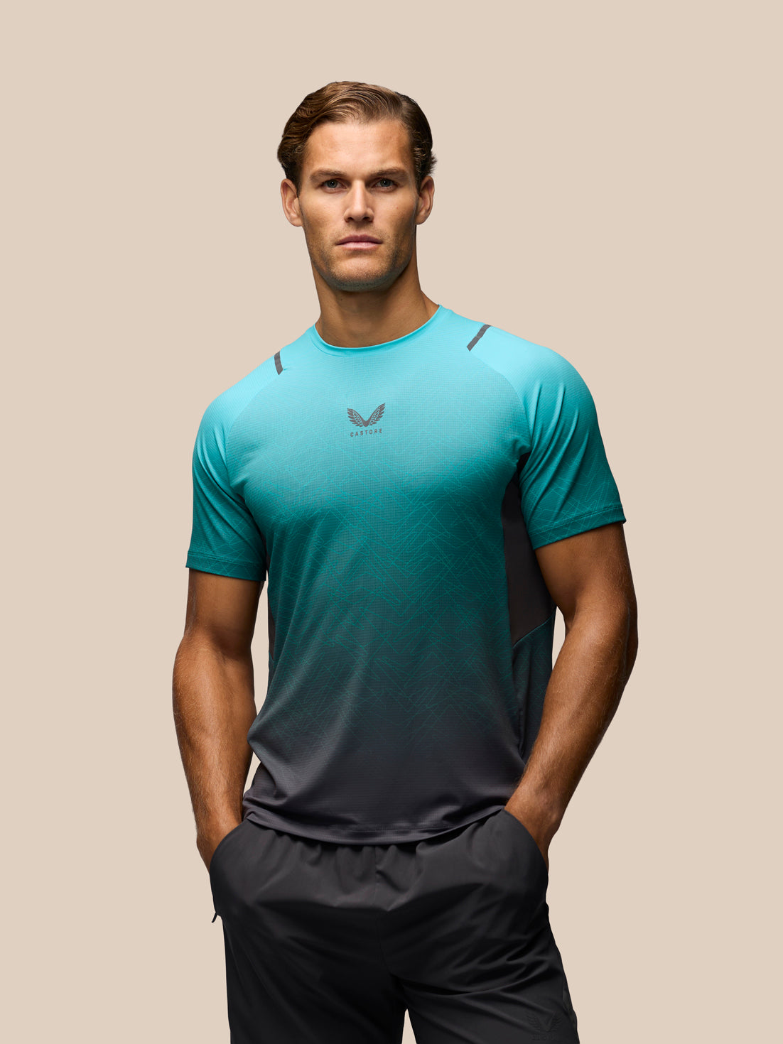 A man wearing a teal and dark gray gradient athletic t-shirt with a small wing logo on the chest, paired with dark gray athletic shorts, set against a solid beige background.