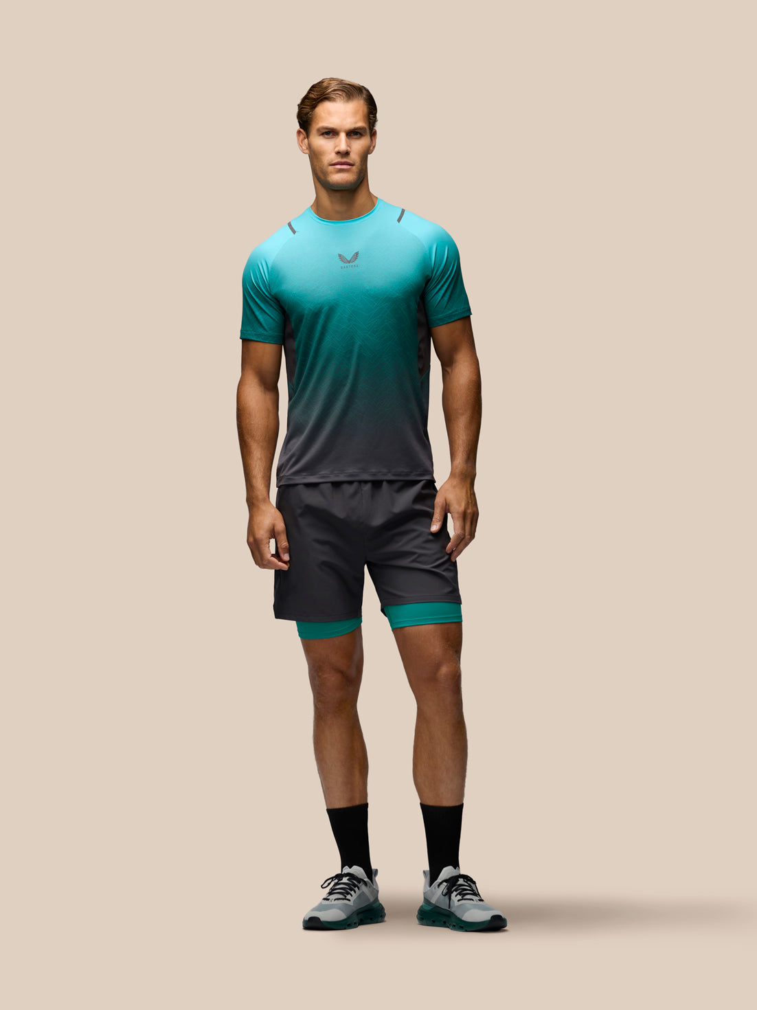 A male model stands against a solid beige background wearing a teal-to-black gradient athletic t-shirt, matching dark gray athletic shorts with a teal inner layer, black socks, and gray running shoes.