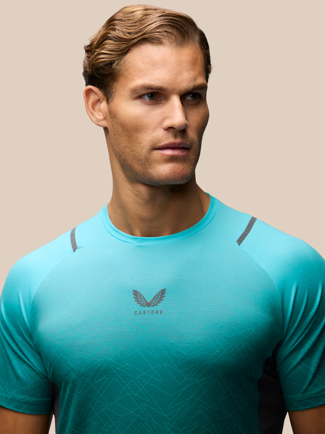 A man wearing a vibrant turquoise athletic t-shirt with a dark gray winged logo and the brand name 'CASTORE' printed on the chest, set against a solid beige background.