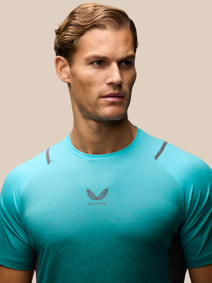 A man wearing a vibrant turquoise athletic t-shirt with a dark gray winged logo and the brand name 'CASTORE' printed on the chest, set against a solid beige background.