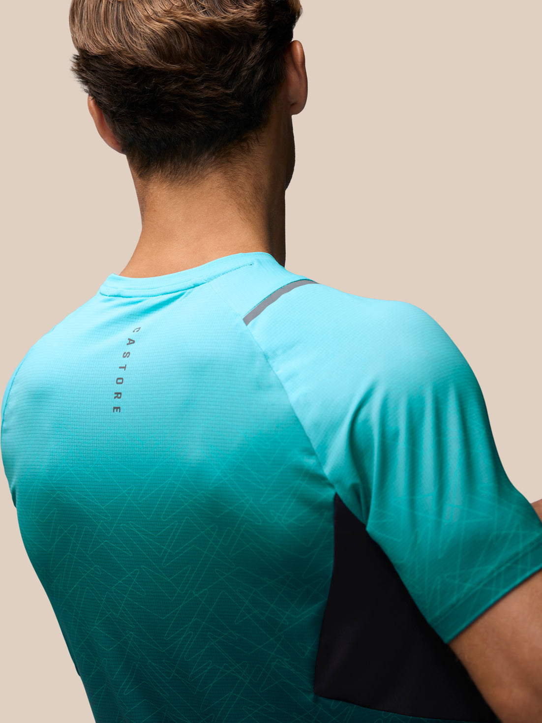 Rear view of a man wearing a turquoise athletic t-shirt with a black side panel and gray shoulder detail, against a solid beige background.
