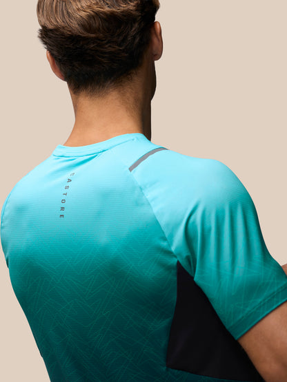 Rear view of a man wearing a turquoise athletic t-shirt with a black side panel and gray shoulder detail, against a solid beige background.