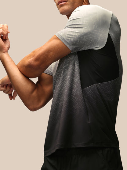 A close-up of a man wearing a short-sleeve athletic shirt with a color-blocked design in light gray, black, and dark gray geometric patterned fabric, shown from the side against a solid beige background.