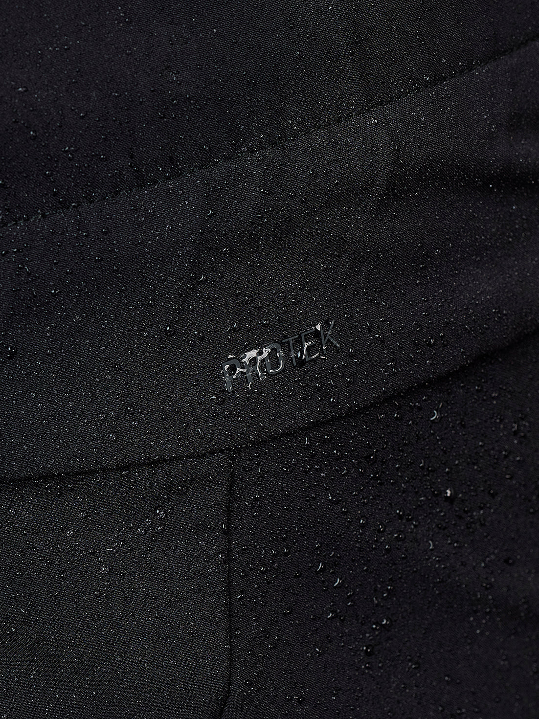 Close-up of a black waterproof fabric with water droplets beaded on the surface and a small metallic 'PROTEK' logo embossed on the seam.