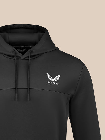 A close-up of a black pullover hoodie with a white winged logo and the word 'CASTORE' on the left chest, shown against a solid beige background.