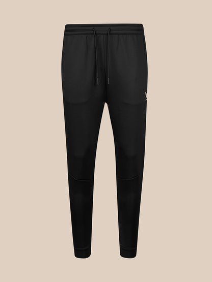 Black jogger-style sweatpants with an elastic waistband, black drawstring, and a small white logo on the left hip, shown against a solid beige background.