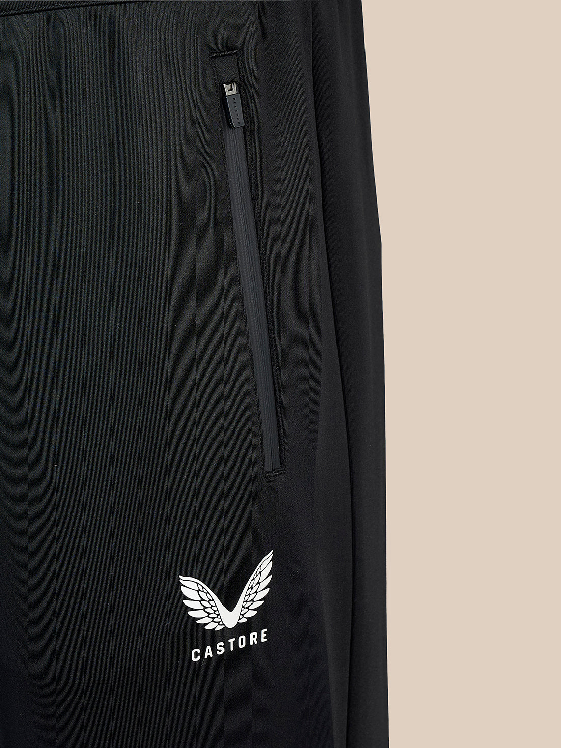 Close-up of black Castore pants featuring a vertical zippered pocket and a white embroidered wing logo with the brand name 'CASTORE' on the lower left leg, set against a solid beige background.