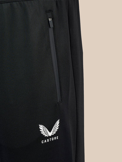 Close-up of black Castore pants featuring a vertical zippered pocket and a white embroidered wing logo with the brand name 'CASTORE' on the lower left leg, set against a solid beige background.