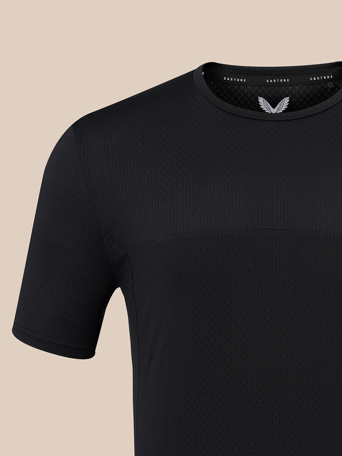 Close-up of a black short-sleeve athletic t-shirt with a textured knit pattern and a small white wing logo on the inside neck label, set against a solid beige background.