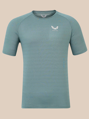 A short-sleeve athletic t-shirt in a muted teal-blue color, featuring a white winged logo and the brand name 'Castore' on the left chest, displayed against a solid beige background.
