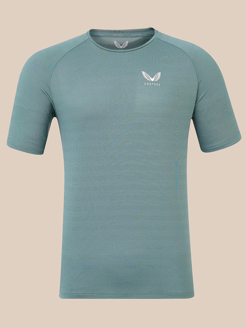 A short-sleeve athletic t-shirt in a muted teal-blue color, featuring a white winged logo and the brand name 'Castore' on the left chest, displayed against a solid beige background.