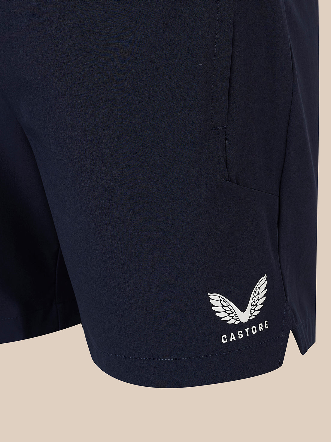 Close-up of navy athletic shorts with a white embroidered winged logo and the word CASTORE near the lower hem, shown against a plain beige background.