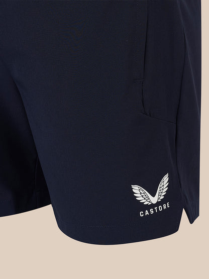 Close-up of navy athletic shorts with a white embroidered winged logo and the word CASTORE near the lower hem, shown against a plain beige background.