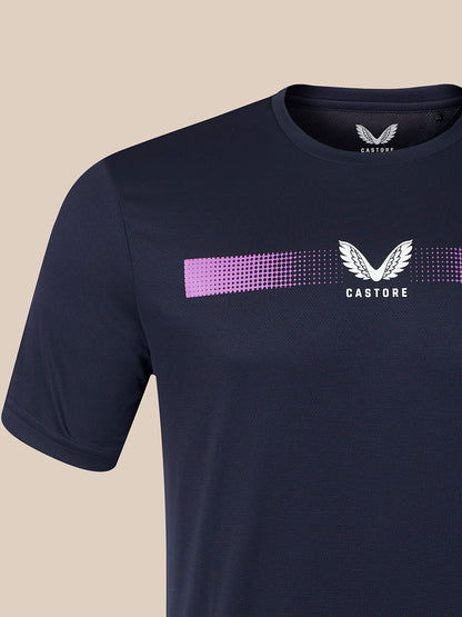 A close-up of a navy blue Castore short-sleeve t-shirt featuring a horizontal dotted purple stripe and a white winged logo with the brand name 'CASTORE' printed on the chest, set against a solid beige background.