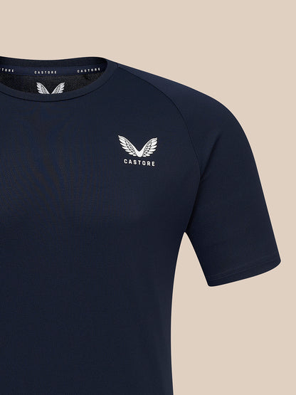 A close-up of a navy blue short-sleeve athletic t-shirt with a white winged logo and the brand name 'CASTORE' printed on the left chest, set against a solid beige background.